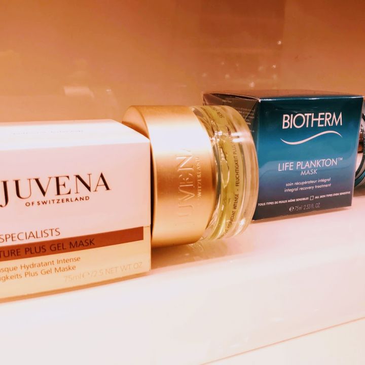 Ein Regal mit Hautpflegeprodukten, darunter die Moisture Plus Gel Mask von Juvena of Switzerland und die Life Plankton Mask von Biotherm, die in einer Reihe angeordnet sind. Die Verpackung ist in den Farben Gold und Blau gehalten.