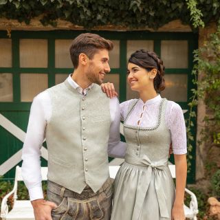 Ein Paar in traditioneller Kleidung steht vor einer rustikalen grün-weißen Tür. Der Mann trägt eine helle Weste und bestickte Hosen, während die Frau ein Kleid mit passender Weste und Schürze trägt. Sie lächeln sich in einer bezaubernden Umgebung im Freien an.