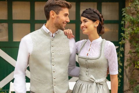 Ein Paar in traditioneller Kleidung steht vor einer rustikalen grün-weißen Tür. Der Mann trägt eine helle Weste und bestickte Hosen, während die Frau ein Kleid mit passender Weste und Schürze trägt. Sie lächeln sich in einer bezaubernden Umgebung im Freien an.