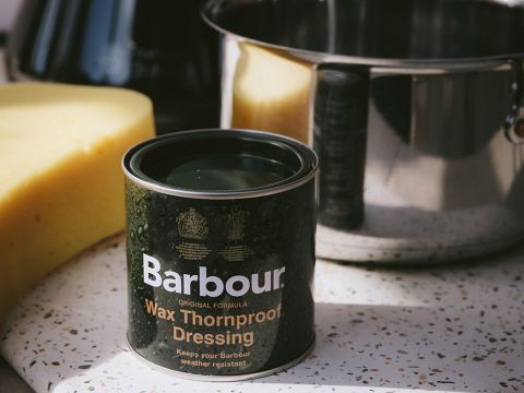 Eine Dose Barbour Wax Thornproof Dressing steht auf einer gesprenkelten Oberfläche neben einem großen gelben Schwamm und einem glänzenden Metalltopf. Die Dose ist dunkel mit weißem und goldenem Text und wird zum Wetterfestmachen von Kleidungsstücken verwendet.