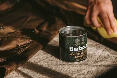 Eine Dose Barbour Wax Thornproof Dressing steht neben einem braunen Stoff oder Kleidungsstück auf einem Tisch. Man sieht eine Person, die das Wachs mit einem gelben Schwamm aufträgt. Die Szene ist warm beleuchtet und schafft eine gemütliche Atmosphäre.