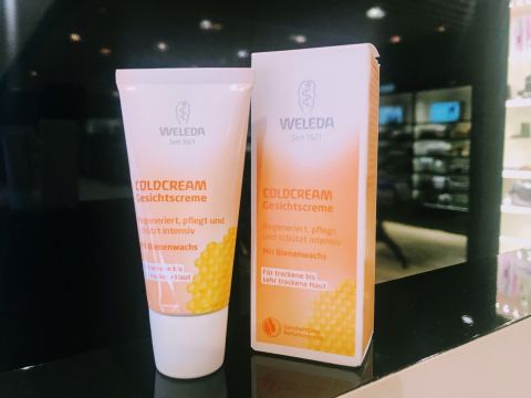 Eine Tube und eine Packung Weleda Coldcream werden ausgestellt. Die Verpackung ist mit Wabenmustern versehen und weist auf deutschen Text hin, der die Vorteile für die Haut und den Bienenwachsgehalt hervorhebt. Der Hintergrund ist ein sanft beleuchtetes Ladenregal.