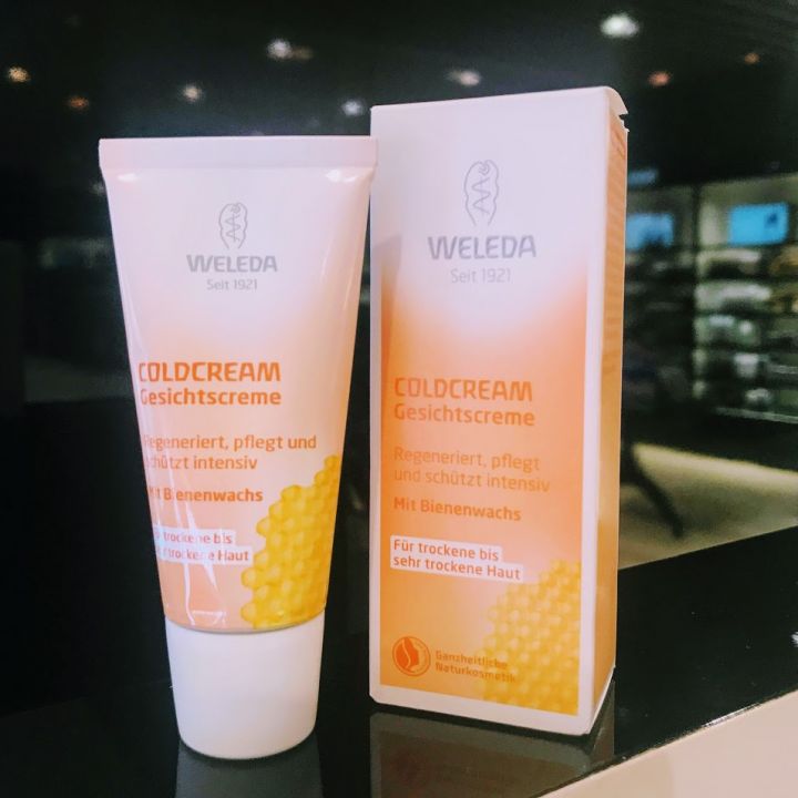 Eine Tube und eine Packung Weleda Coldcream werden ausgestellt. Die Verpackung ist mit Wabenmustern versehen und weist auf deutschen Text hin, der die Vorteile für die Haut und den Bienenwachsgehalt hervorhebt. Der Hintergrund ist ein sanft beleuchtetes Ladenregal.