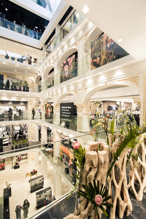 Ein geräumiges, mehrstöckiges Einkaufszentrum mit eleganter Inneneinrichtung, großen Wandgemälden und modernem Design. Es verfügt über Glasgeländer und verschiedene Pflanzen und präsentiert auf mehreren Etagen Bekleidungsgeschäfte und lebendige künstlerische Ausstellungen.