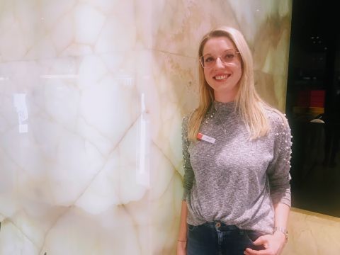 Eine Frau mit langen blonden Haaren und Brille steht lächelnd vor einer von hinten beleuchteten, durchscheinenden Steinwand. Sie trägt einen grauen Pullover mit Perlendetails und Jeans.