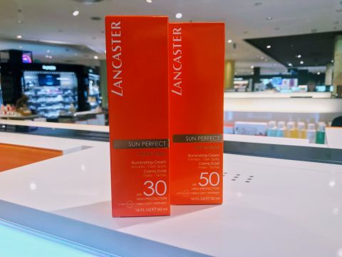 Zwei orangefarbene Schachteln Lancaster Sun Perfect Illuminating Cream mit LSF 30 und 50 stehen auf einem weißen Tresen in einem hell erleuchteten Geschäft. Im Hintergrund sind Regale und andere Produkte zu sehen.
