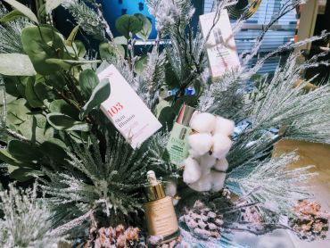 Ein dekoratives Winterarrangement besteht aus bereiftem grünem Laub, Baumwollbüscheln und Tannenzapfen. Zwischen dem Grünzeug befinden sich drei Kosmetikprodukte, darunter Foundation- und Hautpflegeprodukte mit sichtbaren Etiketten. Im Hintergrund sind Regale und verschwommene Ladenbeleuchtung zu sehen.