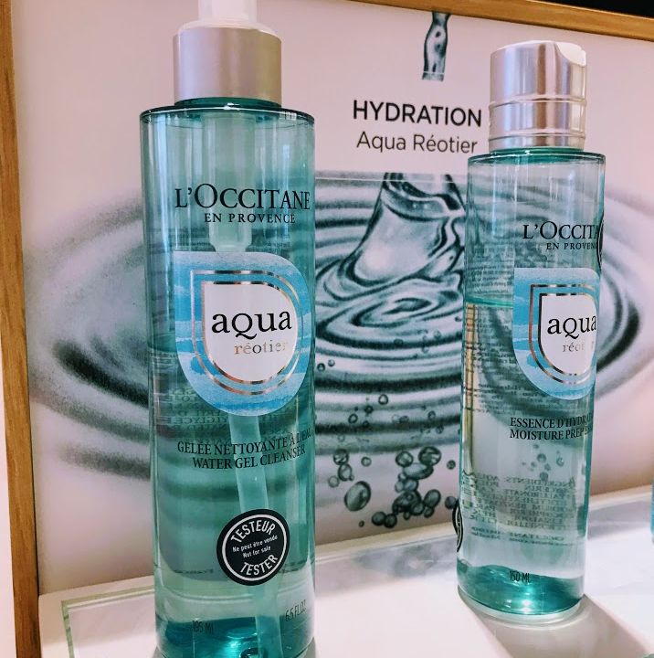 Es werden zwei Flaschen mit Hautpflegeprodukten von L’Occitane Aqua Réotier ausgestellt. Die vordere Flasche ist ein Gelreiniger mit Pumpe und die hintere eine Feuchtigkeitsessenz. Im Hintergrund sind ein Wasserwellenbild und das Wort „Hydration“ zu sehen.