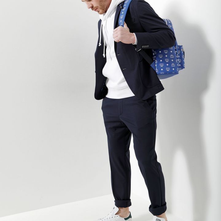 Ein Mann steht vor einer weißen Wand. Er trägt einen marineblauen Anzug, einen weißen Kapuzenpullover und lässige weiße Turnschuhe. Über der Schulter trägt er einen blau gemusterten Rucksack und blickt nachdenklich nach unten.