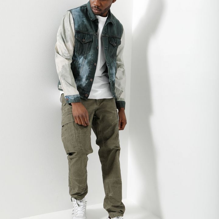 Eine Person steht vor einer weißen Wand. Sie trägt eine Jeansjacke mit Batikmuster, ein weißes T-Shirt, olivfarbene Cargohosen und weiße Turnschuhe. Sie hat einen nachdenklichen Gesichtsausdruck und blickt nach unten. Die Szene ist gut beleuchtet und wirft einen Schatten auf die Wand.