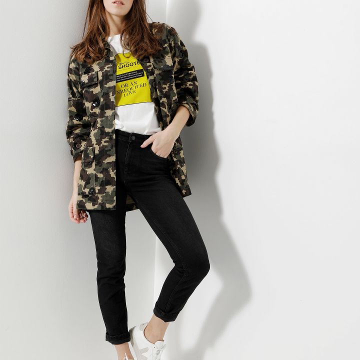 Eine Frau steht vor einer hellen Wand. Sie trägt eine Tarnjacke über einem weißen T-Shirt mit gelbem Text, schwarze Jeans und weiße Turnschuhe. Sie hat lange braune Haare und ihre Hände sind in den Taschen.