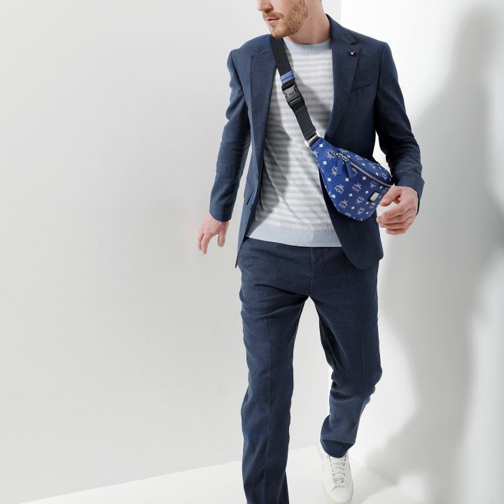 Ein Mann in blauem Anzug und gestreiftem Hemd geht selbstbewusst, trägt weiße Turnschuhe und eine blaue Umhängetasche mit Mustern. Der Hintergrund ist weiß und wirft einen Schatten des Mannes.