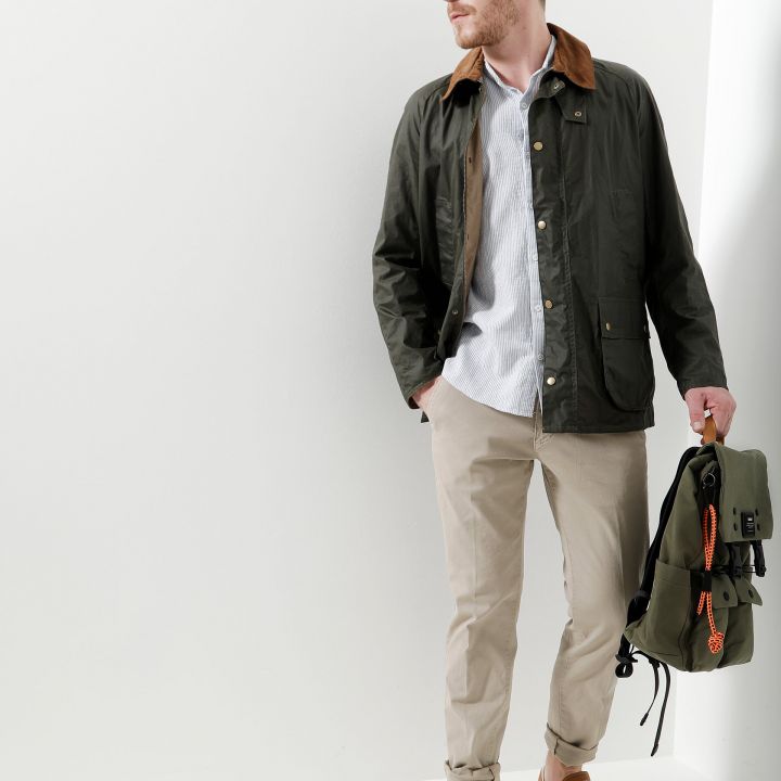 Ein Mann mit kurzen Haaren, einer grünen Jacke, einem gestreiften Hemd, einer beigen Hose und weißen Turnschuhen steht vor einer weißen Wand. In einer Hand hält er einen grünen Rucksack und blickt zur Seite.