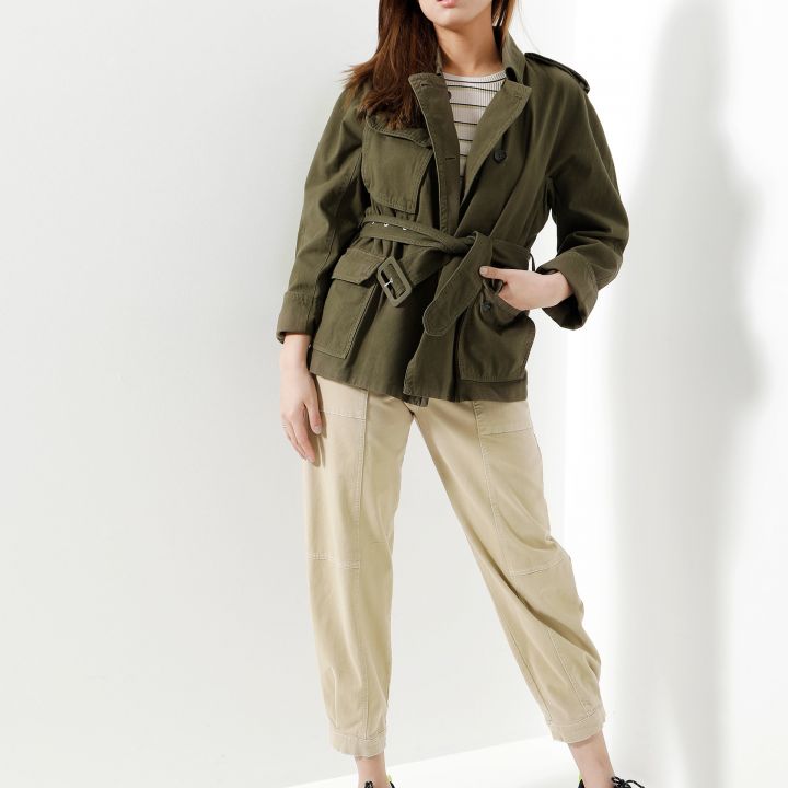 Eine Frau steht selbstbewusst vor einem weißen Hintergrund. Sie trägt eine grüne Arbeitsjacke über einem gestreiften Hemd, beige Hosen und schwarze Turnschuhe mit neongrünen Details. Ihre Hände sind in den Taschen und sie neigt ihren Kopf leicht zur Seite.