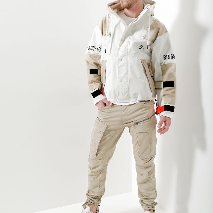 Ein Mann in einer schicken beige-weißen Jacke mit Textdetails auf den Ärmeln steht mit Blick nach links. Er kombiniert die Jacke mit beigen Cargohosen und weißen Turnschuhen. Der Hintergrund ist in einem sanften Weiß gehalten und die Beleuchtung erzeugt eine klare Reflexion.