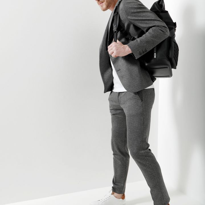 Ein Mann in einem graukarierten Anzug und weißen Turnschuhen steht vor einem weißen Hintergrund. Er trägt einen schwarzen Rucksack über der Schulter und ein weißes T-Shirt.