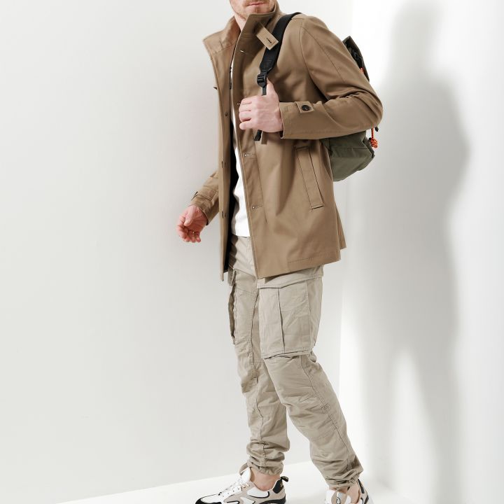 Ein Mann in beiger Jacke und Cargohose steht vor einer weißen Wand. Er trägt einen Rucksack und schaut zur Seite. Er hat kurze Haare und einen Bart und trägt weiße und graue Turnschuhe.
