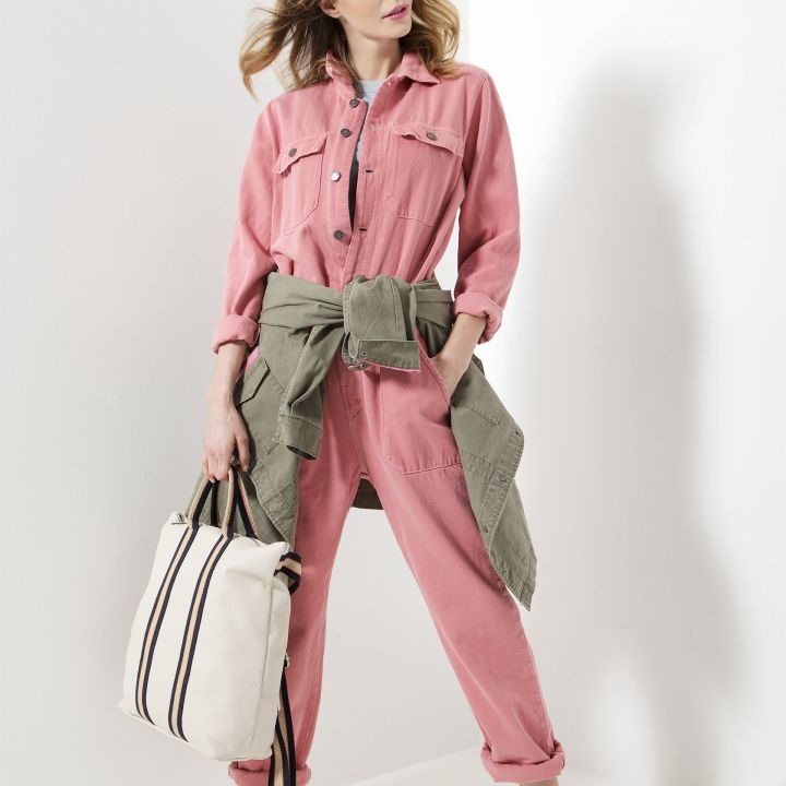 Eine Frau steht selbstbewusst in einem rosa Overall mit einer Khakijacke um die Hüfte. Sie hält eine weiß-schwarz gestreifte Tasche in der Hand und trägt weiße Turnschuhe. Ihr Haar ist sanft gewellt und sie steht vor einer weißen Wand.