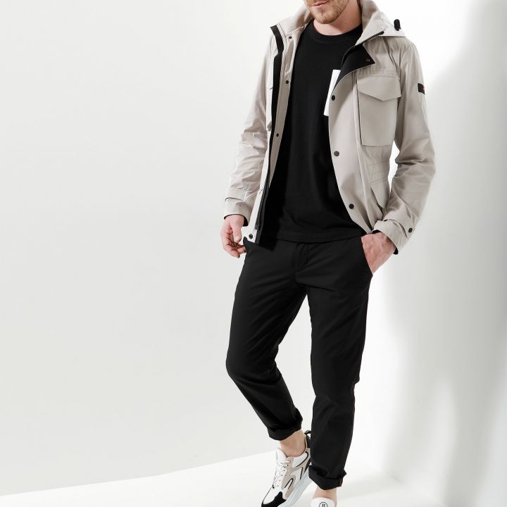 Ein Mann steht vor einem weißen Hintergrund. Er trägt eine hellgraue Jacke über einem schwarzen Hemd, eine schwarze Hose und weiße Turnschuhe mit schwarzen und braunen Akzenten.