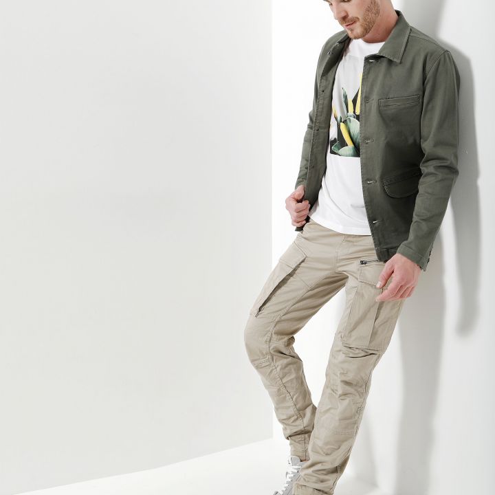 Ein Mann mit kurzen Haaren lehnt an einer weißen Wand. Er trägt eine grüne Jacke, ein T-Shirt mit Aufdruck, beige Cargohosen und weiße Turnschuhe. Seine Hände stecken in den Hosentaschen und er schaut nach unten. Die Umgebung ist hell erleuchtet und minimalistisch gestaltet.