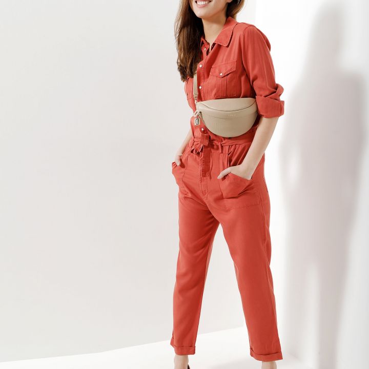 Eine Frau in einem roten Overall und hellbraunen Slippern steht lächelnd mit den Händen in den Taschen da. Sie trägt eine beige Gürteltasche und steht vor einem schlichten, hellen Hintergrund. Ihr langes Haar ist zur Seite frisiert.