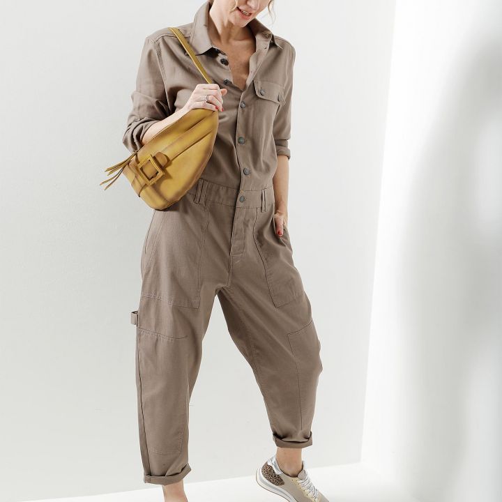 Eine Person in einem braunen Overall mit hochgekrempelten Ärmeln und Hosenbeinen steht vor einem weißen Hintergrund. Sie trägt eine senfgelbe Handtasche und beige Turnschuhe. Die Hände stecken in den Taschen und die Haare sind zurückgebunden.