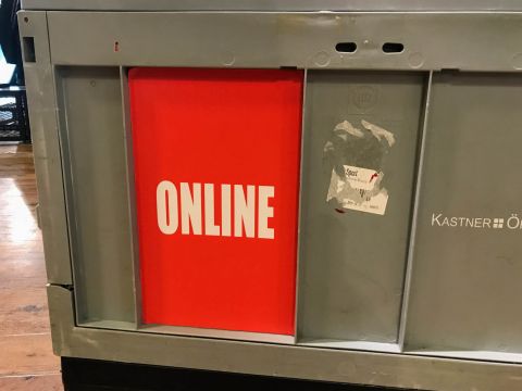 Eine Nahaufnahme einer grauen Tafel mit einem roten Abschnitt, auf dem in fetten weißen Buchstaben das Wort „ONLINE“ steht. Die Tafel enthält ein Etikett mit dem Text „KASTNER ÖHL“ und einen abgenutzten Aufkleber. Im Hintergrund ist ein Holzboden zu sehen.