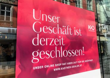 Auf einem Schaufensterschild mit rotem Hintergrund steht: „Unser Geschäft ist derzeit geschlossen! Unser Online-Shop hat aber 24/7 für Sie geöffnet! www.kastner-oehler.at“ und weist darauf hin, dass das Ladengeschäft geschlossen, der Online-Shop jedoch geöffnet ist.