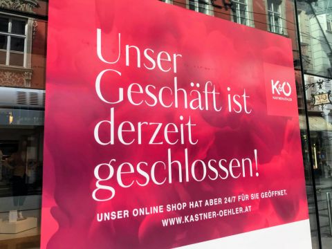 Auf einem Schaufensterschild mit rotem Hintergrund steht: „Unser Geschäft ist derzeit geschlossen! Unser Online-Shop hat aber 24/7 für Sie geöffnet! www.kastner-oehler.at“ und weist darauf hin, dass das Ladengeschäft geschlossen, der Online-Shop jedoch geöffnet ist.