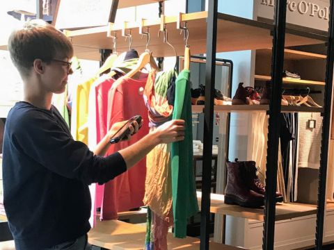 Eine Person mit Brille steht in einem Bekleidungsgeschäft und scannt mit einem Smartphone ein grünes Hemd, das zwischen verschiedenen bunten Kleidungsstücken auf einem Ständer hängt. In den Regalen daneben sind Schuhe und Accessoires ausgestellt.