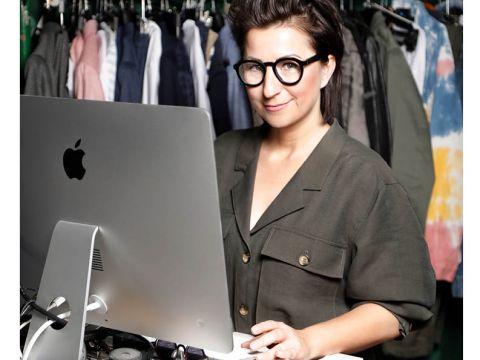 Eine Person mit kurzen Haaren und runder Brille steht an einem Schreibtisch mit einem großen Monitor. Sie trägt ein grünes Hemd mit Knöpfen. Hinter ihr hängen Kleider auf einem Ständer.
