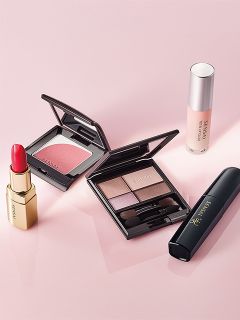 Eine Reihe von Make-up-Produkten, darunter Lippenstift, Rouge, Lidschattenpalette, Wimperntusche und Grundierung, die auf einer rosa Fläche angeordnet sind.