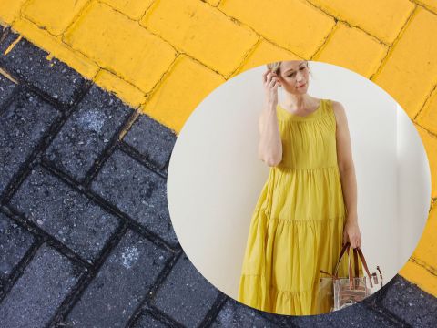 Eine Person in einem fließenden gelben Kleid hält eine durchsichtige Handtasche vor einem Hintergrund aus ineinandergreifenden gelb und schwarz gemusterten Gehwegsteinen.