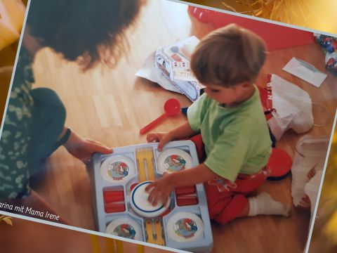Ein Kleinkind mit kurzen braunen Haaren sitzt auf einem Holzboden und spielt mit einem bunten Spielzeugset. Die Hand eines Erwachsenen hilft ihm. Überall liegen Spielzeugteile verstreut herum, außerdem ein roter Löffel und eine offene Schachtel.