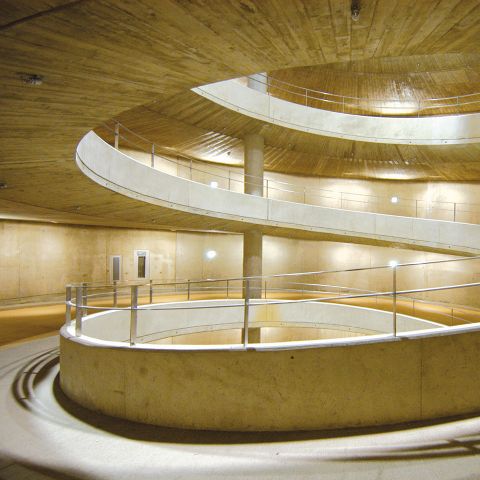 Eine geschwungene, mehrstufige Beton-Spiralrampe im Inneren eines gut beleuchteten runden Gebäudes mit glatten Wänden und Metallgeländern. Das Design vermittelt ein Gefühl von Tiefe und modernem Architekturstil.