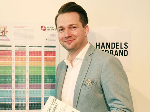 Ein Mann im grauen Anzug hält eine Broschüre in der Hand und steht vor einem Wanddisplay mit dem Logo des Handelsverbandes und bunten Diagrammen.