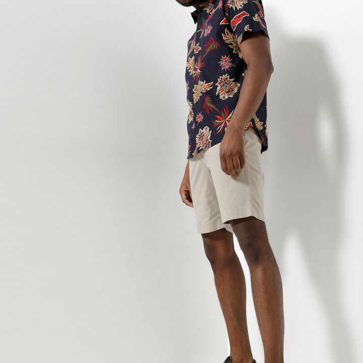Eine Person mit Sonnenbrille, dunklem Kurzarmhemd mit Blumenmuster, hellbeigen Shorts und schwarzen Turnschuhen steht selbstbewusst vor einem schlichten weißen Hintergrund.