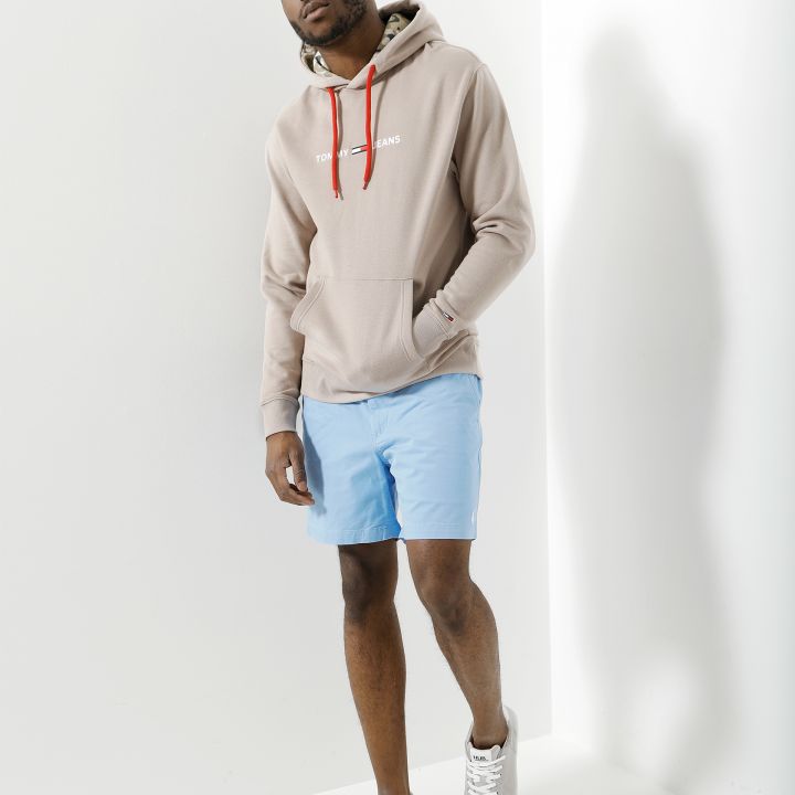 Ein Mann posiert in einem beigen Kapuzenpullover mit roten Kordeln und blauen Shorts. Er trägt graue Turnschuhe und steht in einem gut beleuchteten Raum mit weißen Wänden und weißem Boden.