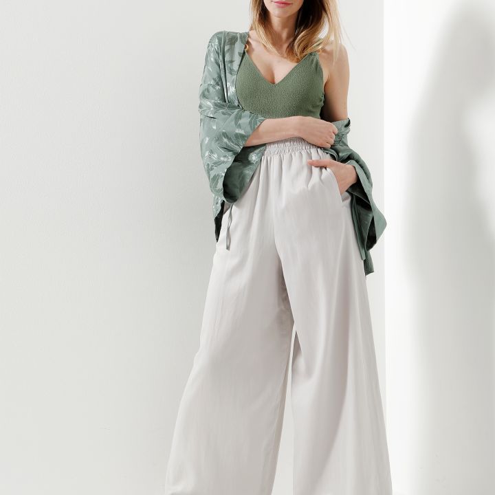 Eine Frau steht in einer minimalistischen Umgebung und trägt ein grünes Oberteil, eine gemusterte offene Strickjacke und weiße Hosen mit weitem Bein. Ihre Hände sind in den Taschen und sie trägt grün-weiße Pantoletten. Sie blickt direkt in die Kamera.