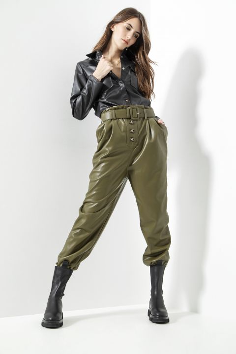 Eine Frau steht vor einem weißen Hintergrund und trägt ein schwarzes Lederhemd und hoch taillierte olivgrüne Lederhosen mit Gürtel. Sie kombiniert das Outfit mit schwarzen Stiefeln, ihr langes Haar fällt herab und sie nimmt eine selbstbewusste Pose ein.