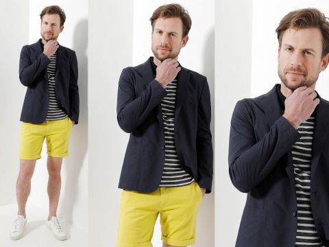 Ein Mann posiert aus drei verschiedenen Winkeln. Er trägt einen marineblauen Blazer über einem gestreiften Hemd und gelben Shorts, dazu weiße Turnschuhe. Er steht vor einem hellen Hintergrund und berührt nachdenklich sein Kinn.