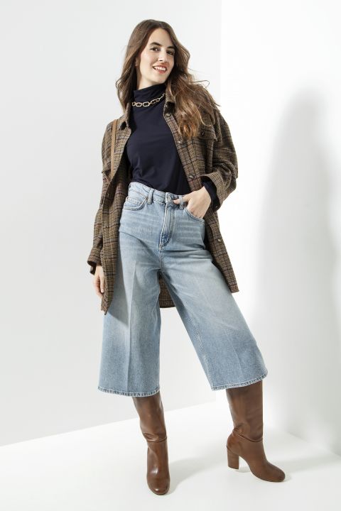 Eine Frau in kariertem Mantel und schwarzem Hemd posiert selbstbewusst. Sie trägt hellblaue Jeans mit weitem Bein und braune kniehohe Stiefel. Der Hintergrund ist minimalistisch und hell und unterstreicht ihr lässiges und doch stilvolles Outfit.
