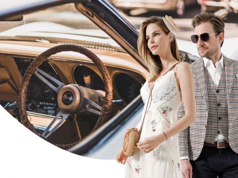 Eine Frau im weißen Kleid und ein Mann im karierten Anzug stehen selbstbewusst neben einem Bild des hölzernen Lenkrads und Armaturenbretts eines Oldtimers. Die Szene vereint Eleganz und klassischen Automobilstil.