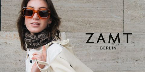Eine Person mit einer großen Sonnenbrille und einem gemusterten Schal posiert vor einer Steinmauer. Über der Schulter trägt sie eine helle Tasche. Neben ihr prangt der Text „ZAMT BERLIN“.