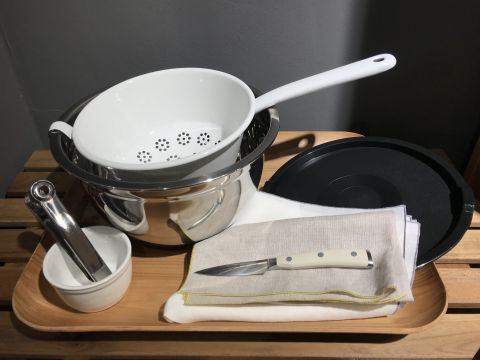Eine Kücheneinrichtung mit einem Edelstahltopf, einem weißen Sieb, einem schwarzen Deckel, einem Mörser und Stößel sowie einem Messer, das auf einer gefalteten Stoffserviette auf einem Holztablett liegt.