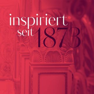Ein rot getöntes Bild mit dekorativen Architekturdetails und dem Text „inspiriert seit 1873“ in weißer und schwarzer Schrift.