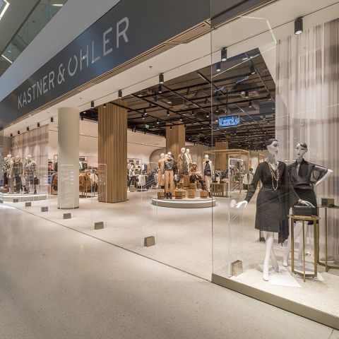 Schaufenster von Kastner & Öhler mit Schaufensterpuppen in modischer Kleidung. Das Innere ist gut sichtbar und zeigt modernes Dekor mit beleuchteten Kleiderständern und Accessoires. Das Geschäft hat Glaswände und ein klares, geräumiges Design.