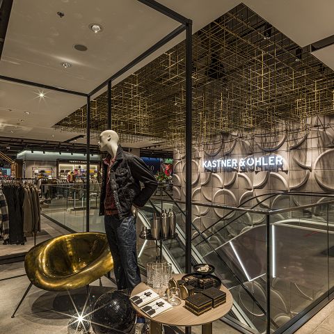 Innenansicht eines modernen Bekleidungsgeschäfts mit einer Schaufensterpuppe in Jeansjacke und Jeans, einem goldenen Stuhl und einem kleinen Tisch mit verschiedenen Dekorationsgegenständen. Die Decke ist geometrisch gestaltet und im Hintergrund ist ein „Kastner & Öhler“-Schild zu sehen.