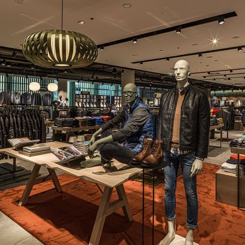Innenansicht eines Bekleidungsgeschäfts mit zwei Schaufensterpuppen in Freizeitkleidung. Eine trägt eine Lederjacke und Jeans, die andere eine Pufferweste und ein Hemd. Auf Regalen sind Kleidungsstücke ausgestellt, und Deckenleuchten sorgen für eine warme, moderne Atmosphäre.