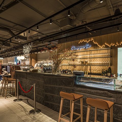 Ein modernes Bar-Interieur mit Steinakzenten und Holzelementen. Die Bar verfügt über ein Neonschild, Glaswarenstapel und Flaschen auf Regalen. Vor der Theke stehen Hocker und Blumendekorationen sorgen für einen Hauch von Farbe.
